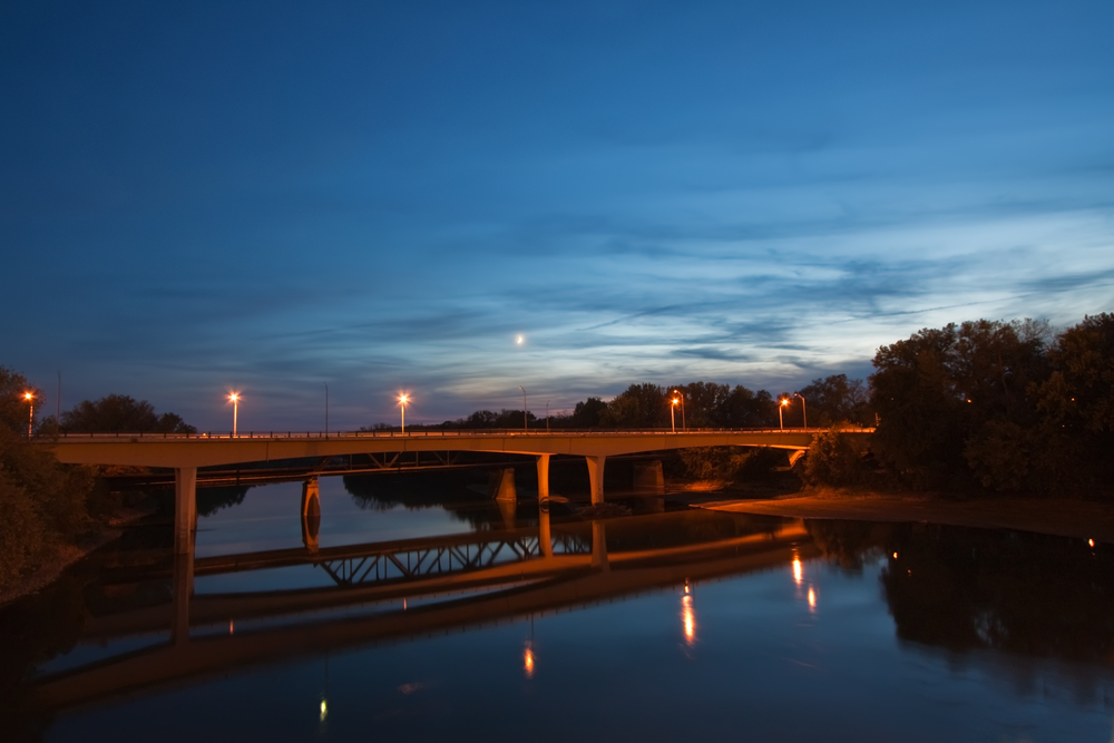 Bridge over the Wabash River near Lafayette, Indiana with dramatic blue sky and a crescent moon as night falls over the city